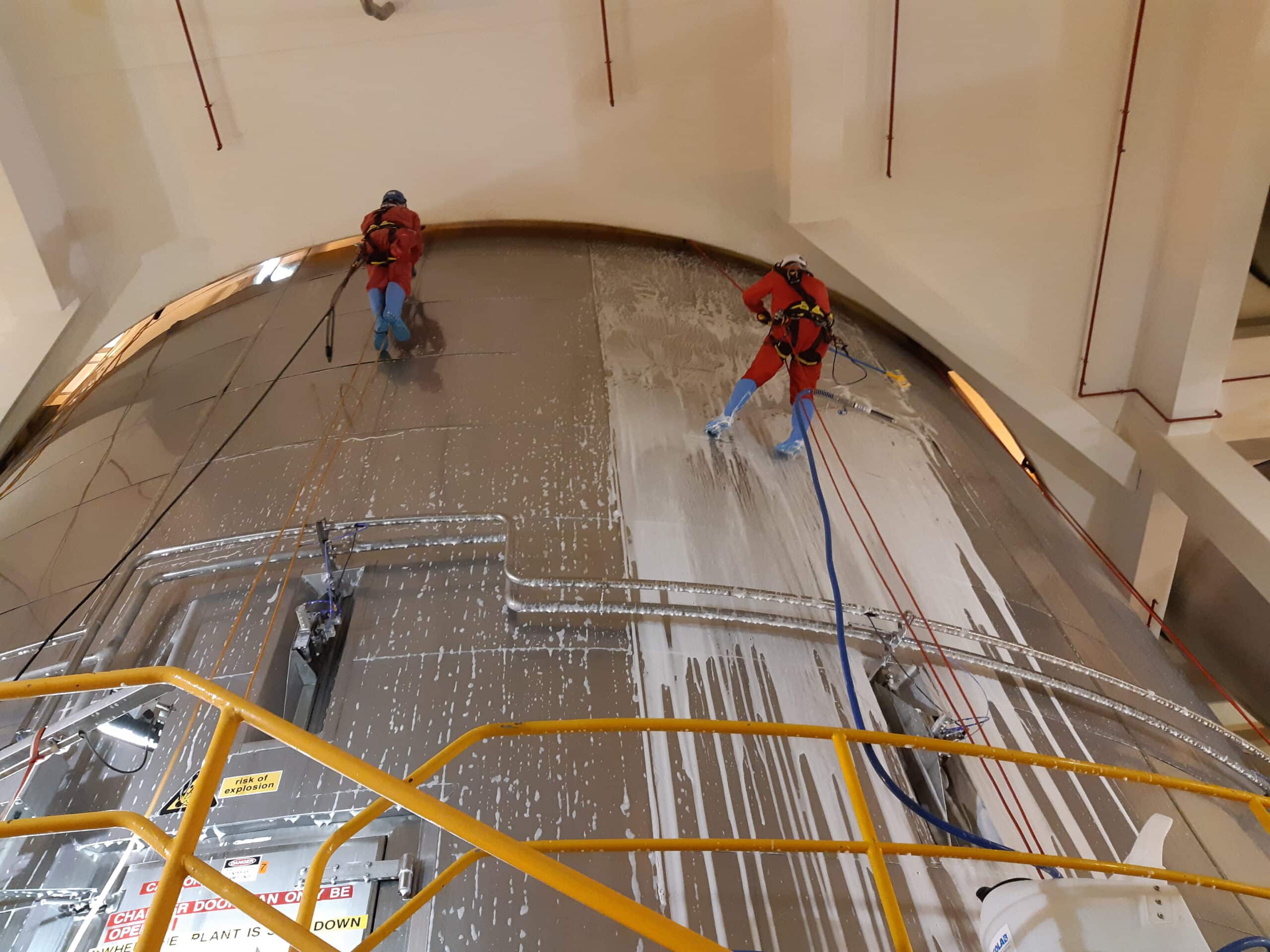 Two Connect Access industrial rope access technicians cleaning the exterior of a large stainless-steel tank at a Fonterra facility, suspended by safety ropes and using industrial abseiling techniques to perform high-level maintenance safely and efficiently.