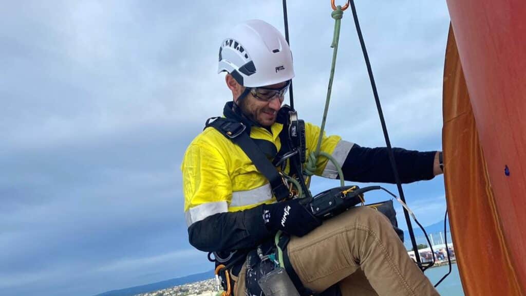 A close-up of industrial abseiler Brendon Kinnaird suspended on rope access lines working on a high structure above a harbour and waterfront road.
