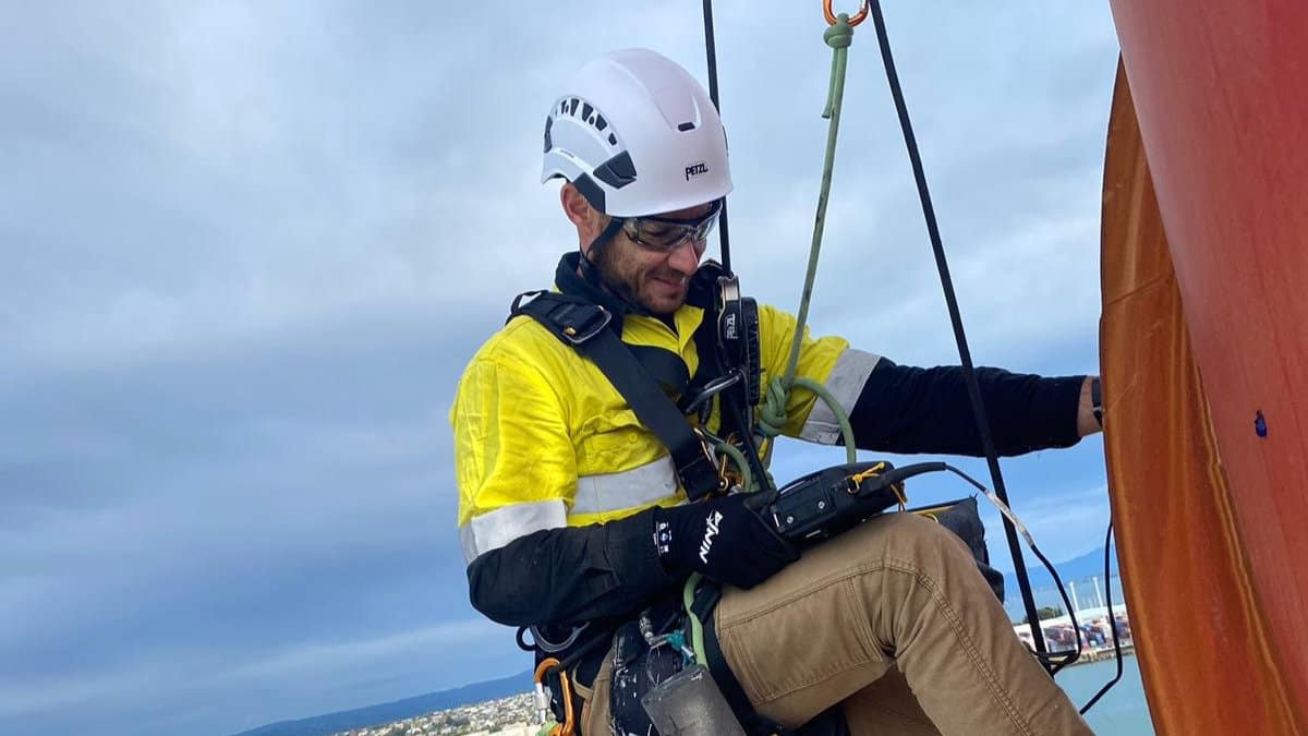 A close-up of industrial abseiler Brendon Kinnaird suspended on rope access lines working on a high structure above a harbour and waterfront road.