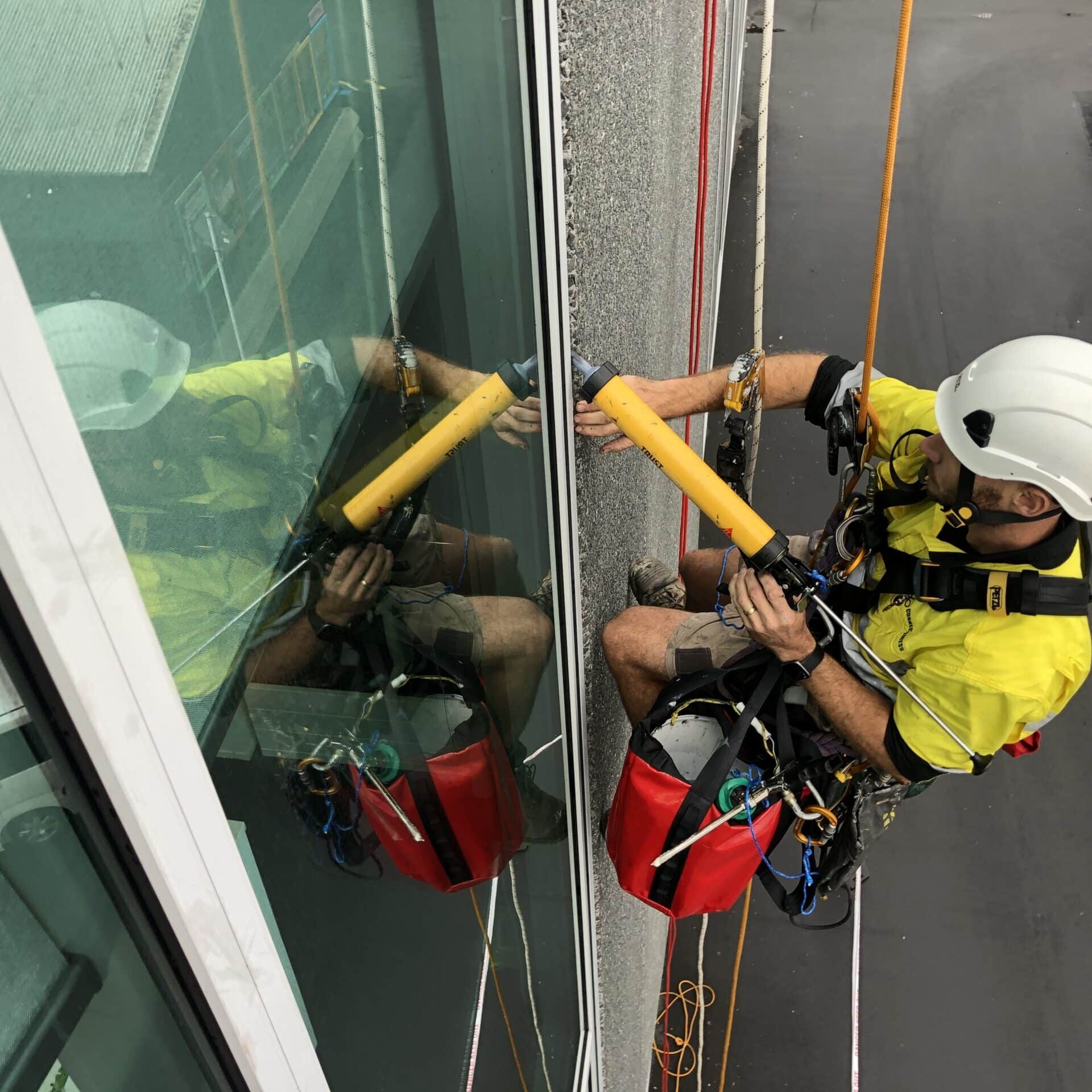 Technician performing rope access expansion joint replacement on a high-rise building, wearing safety gear and working efficiently on the exterior facade